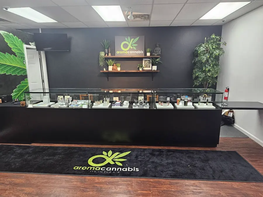 Inside Aroma Cannabis: a black counter with glass display cases, wooden shelves holding plants and the aromacannabis 'It's in the Air' sign, a Buddha statue, and the leaf logo floor mat.